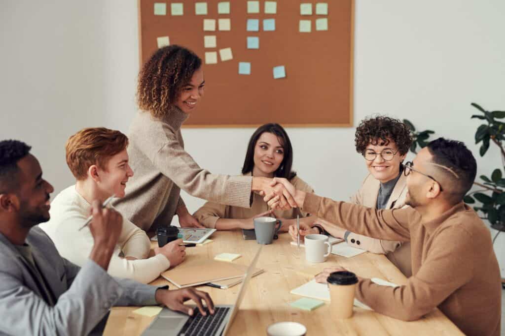 Six people sitting at a desk in a meeting with two people shaking hands