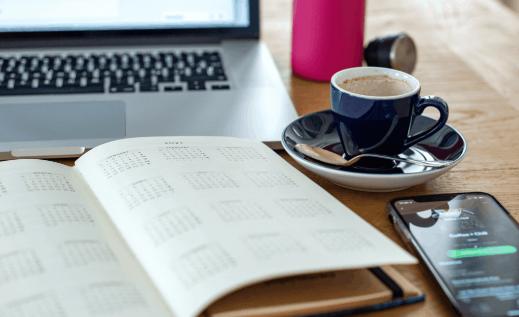 Calendar notebook open on table with laptop, cellphone, and coffee cup