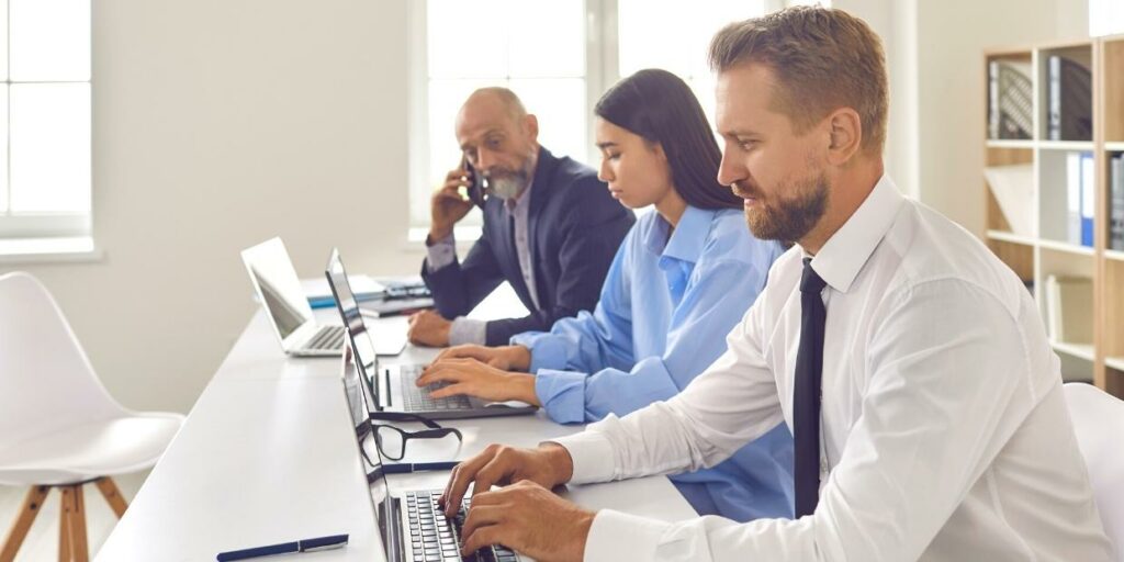 Three coworkers at a desk using laptops and cellphones